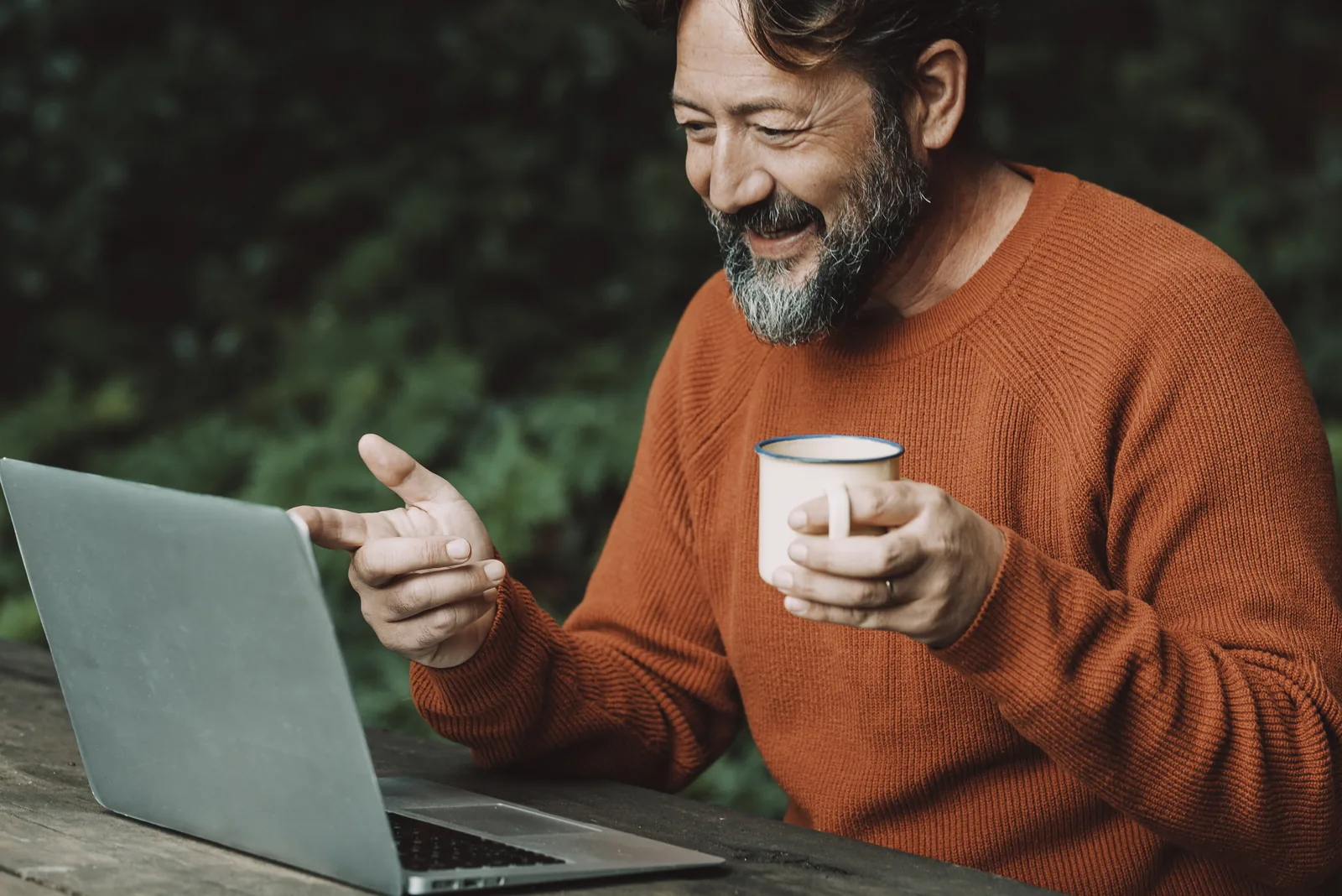 Smiling man holding a coffee mug while using a laptop outdoors