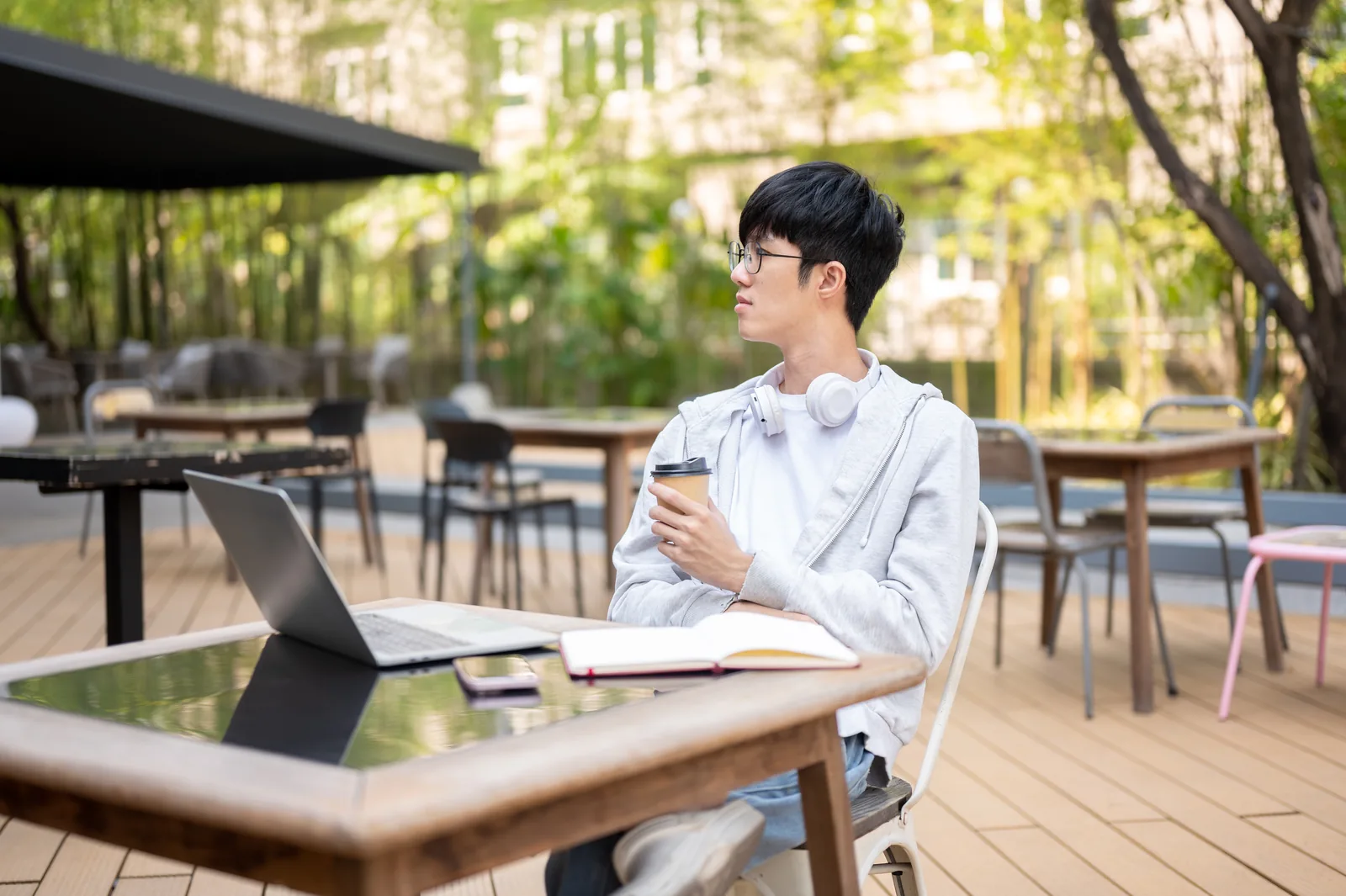 Young man holding coffee beside a laptop and open notebook at an outdoor café