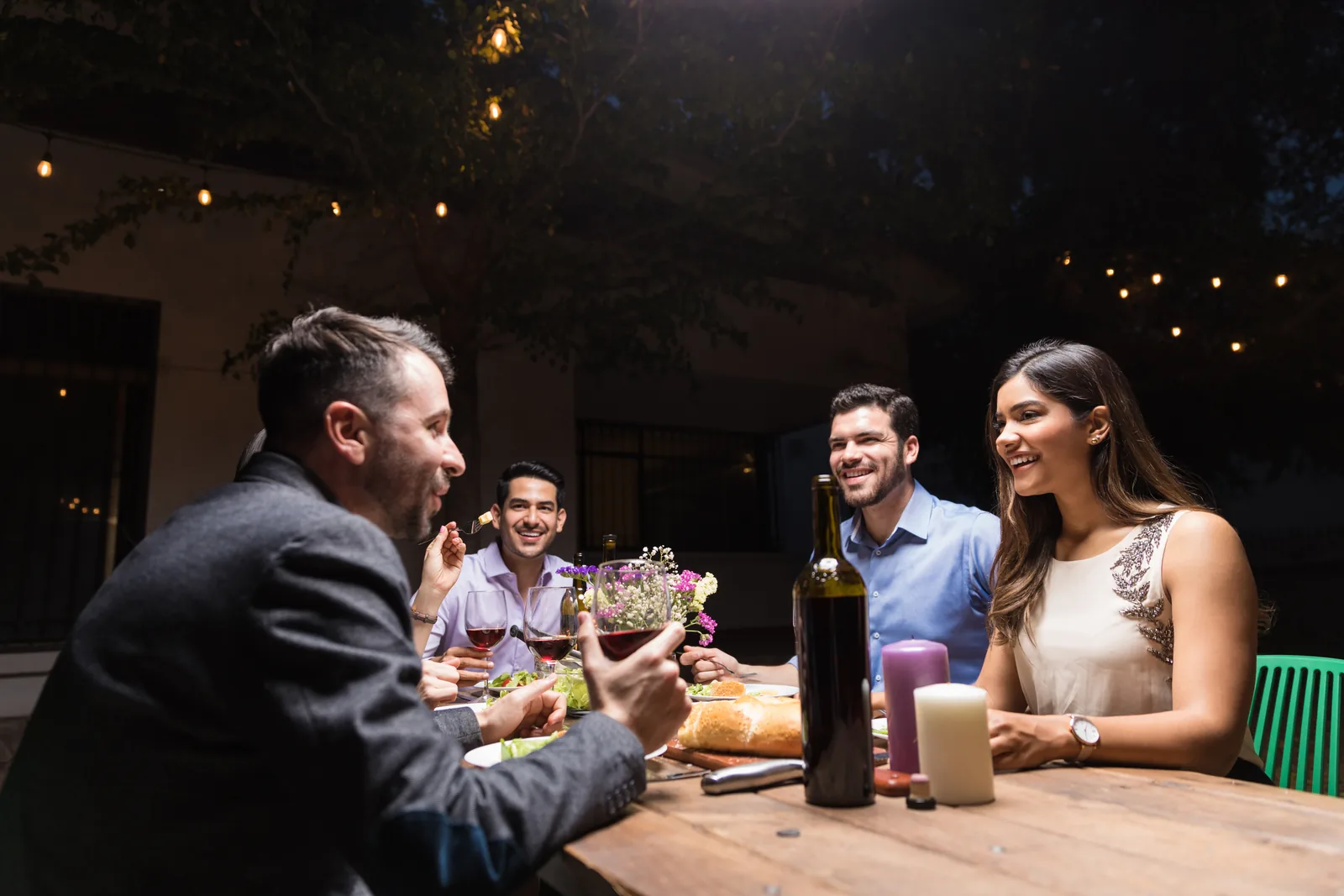 Friends sharing a meal and conversation at an outdoor table beneath string lights