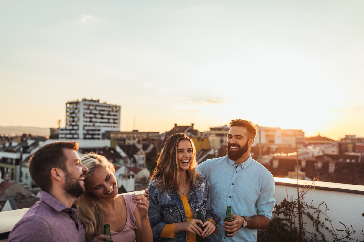 Four friends laughing and enjoying drinks on a rooftop at sunset